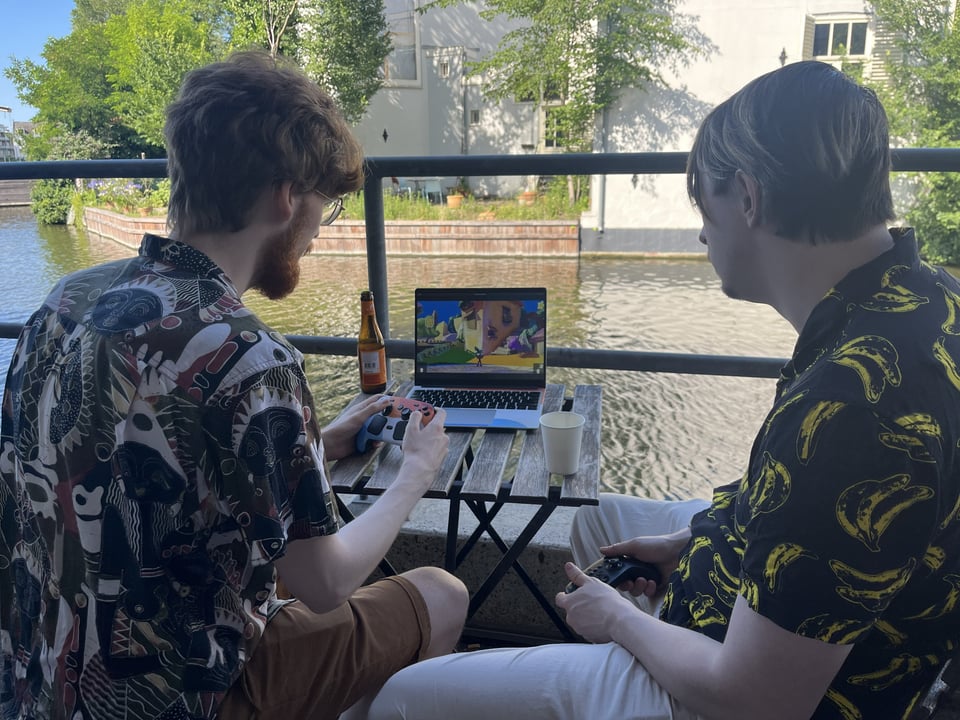 Two people, controllers in hand, play a game on a laptop perched on a canal-side table.
