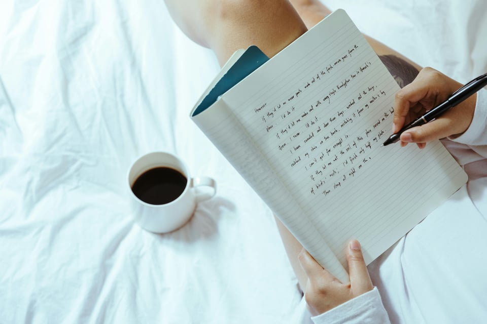 Image of a person writing in a notebook (face not shown) while sitting on a bed or couch with a cup of coffee beside them.