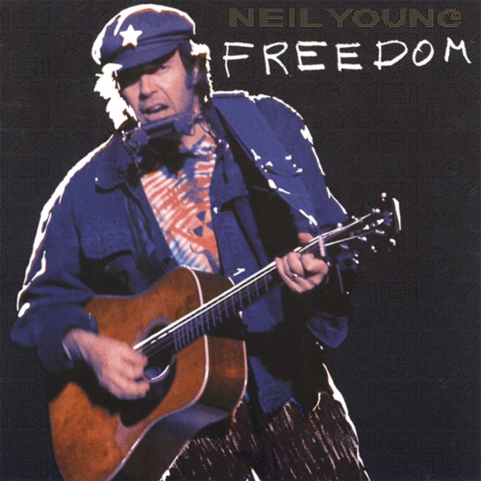 A color photgraph of Neil Young, a white man in a blue jacket, blue cap with a white star on it, and tie-dyed shirt with a radiation symbol on it. He is singing. He exhibits the most "cranky uncle" energy in the universe. He's playing acoustic guitar and has one of those harmonica holders on his neck. It looks like it was shot in concert.