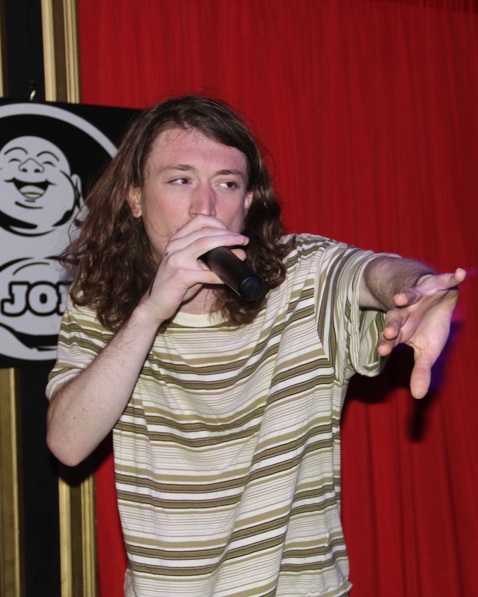 A young man in a striped short-sleeve shirt with shoulder-length light brown hair holds a microphone to his mouth with his right hand, with his left hand stretched toward the camera.