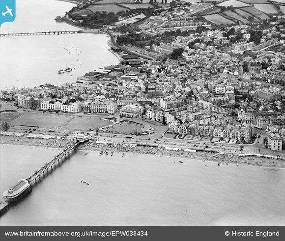 An aerial view of Teignmouth in 1930, looking across the town from the sea. In the foreground is the then larger pier and a large empty green space (the Den). Beyond is the Georgian town and the railway. Beyond that, are fields, one of which has a handful of white houses in it.