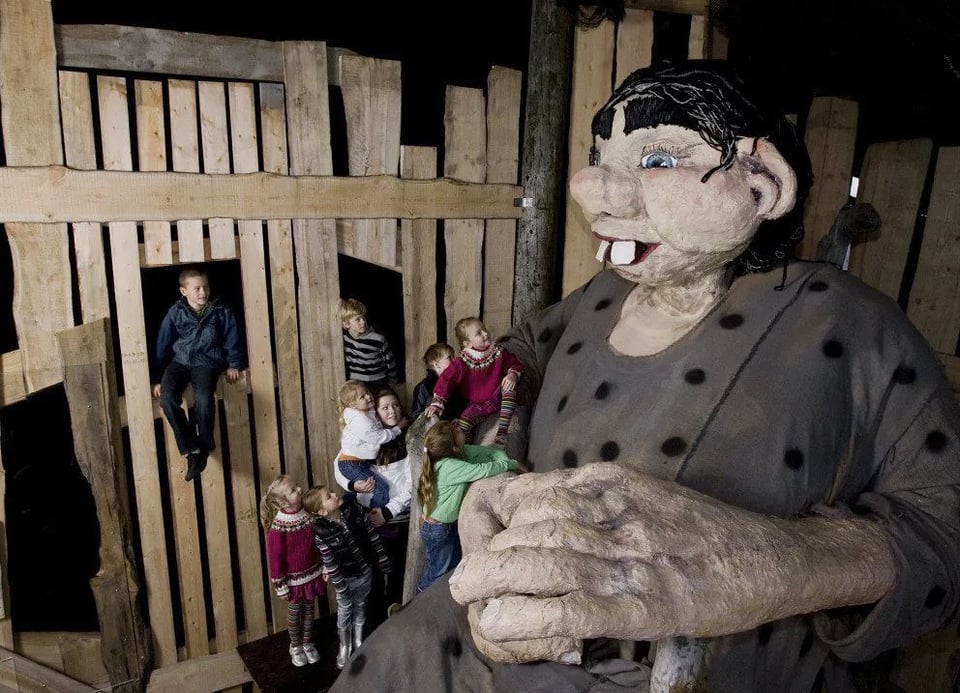 A dozen chlidren climb through a crude wooden wall to look at a gigantic, crude papier-mache giantess with thick eyebrows and buck teeth.
