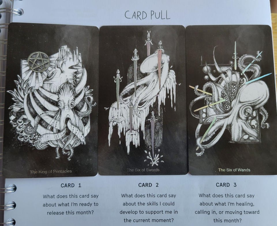 image description: a row of three octopus-themed black and white tarot cards on top of a notebook. The cards have splashes of holographic bits; each card sits above a reflection question printed in the notebook. The first card is the king of pentacles, depicting an octopus with crown-like head decoration at the base of a coral castle with a shiny pentacle in the top left corner. Beneath it the notebook reads ‘What does this card say about what I’m ready to release this month?’ The second card is the 6 of swords, depicting an octopus moving towards the top right corner, weaving between 6 shiny, downward-pointing daggers. Beneath it the notebook reads ‘What does this card say about the skills I could develop to support me in the current moment?’ The third card is the 6 of wands, depicting an octopus emerging from an abstractly-shaped frame holding 6 shiny wands in its arms, loosely radiating out from its center. Beneath it the notebook reads ‘What does this card say about what I’m healing, calling in, or moving towards this month’ end image description.