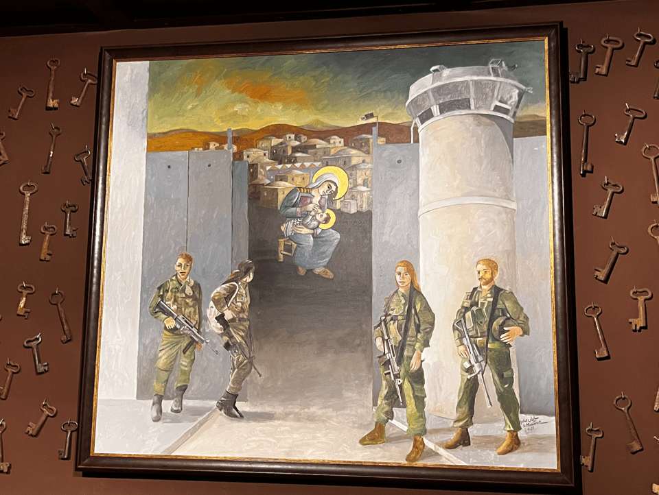 Photograph of a framed painting on a brown background covered with old-fashioned metal keys. The painting shows four soldiers with machine guns in front of a tall concrete wall with a watch tower. Behind an opening in the wall we see a village in the background, along with a seated woman holding a baby. The woman and the baby are painted with yellow halos.