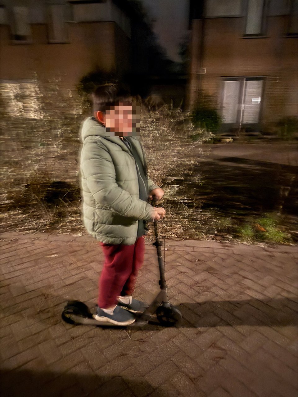 A boy goes along a street on a scooter. He face is blurred, but most of his body is in focus and the street is blurred from camera motion.