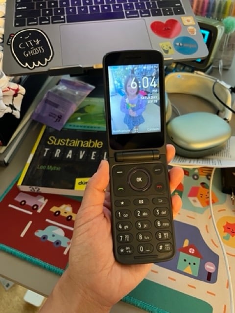 my hand holding a flip phone, opened so that you can see the screen lit up and the number keypad. The phone is displaying the time and is being held above a cluttered desk