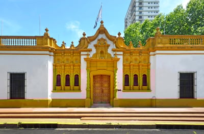 A white building of neo-colonial Hispanic architecture, with mustard yellow paint and a large heavy wooden door.