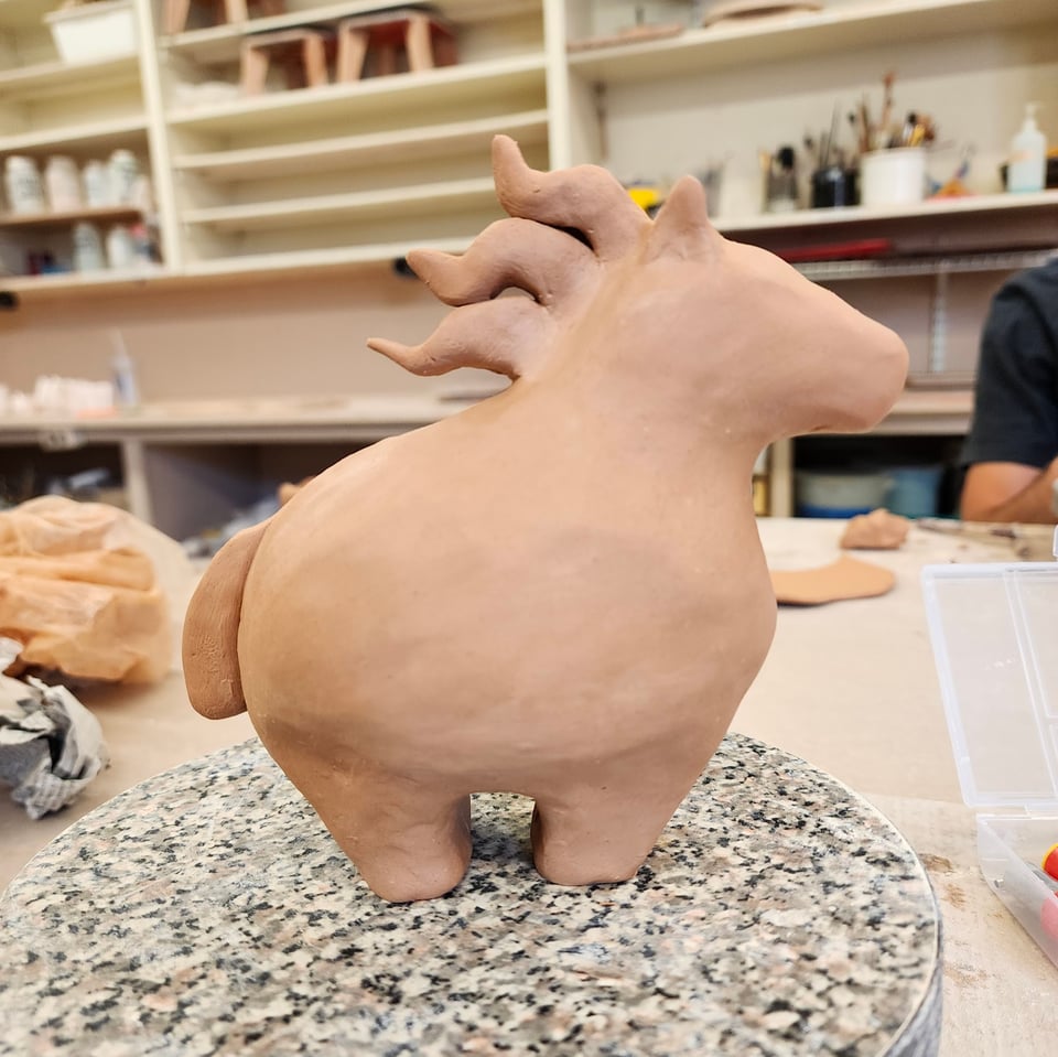 A clay horse sits on a table in an art studio. The horse is round. VERY round. It's mane is three jaunty lightning bolts, its chonky feet barely supporting its chonkitude.