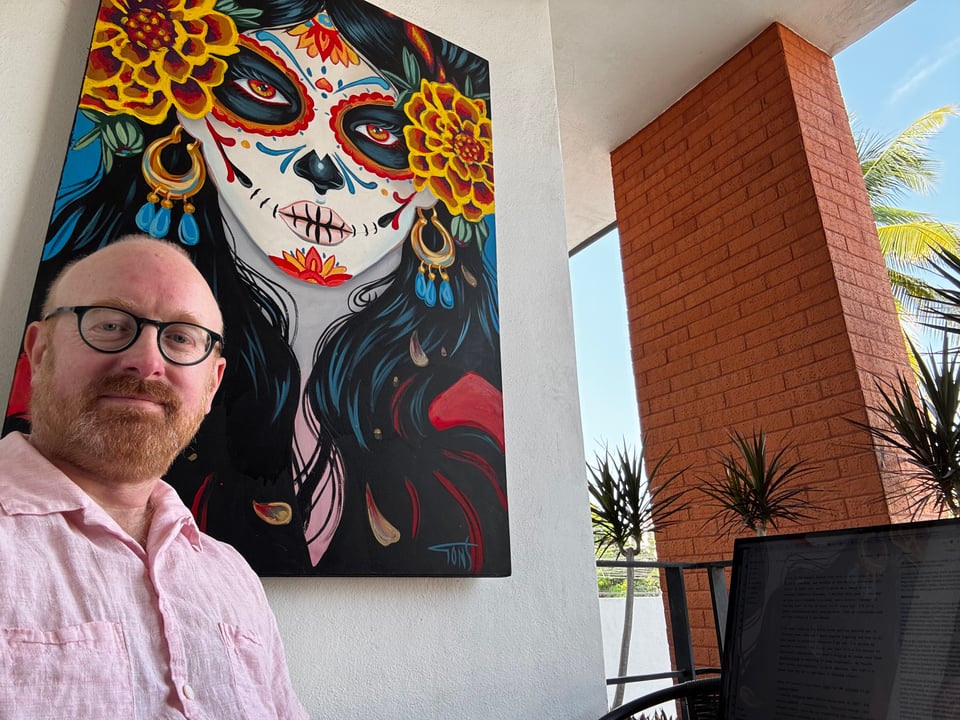Matt is sitting with his laptop on a balcony, smiling in a loose pink button-up. Behind him on the wall is a large painting of the legendary La Catrina, a feminine skeletal figure adorned with paint and marigolds