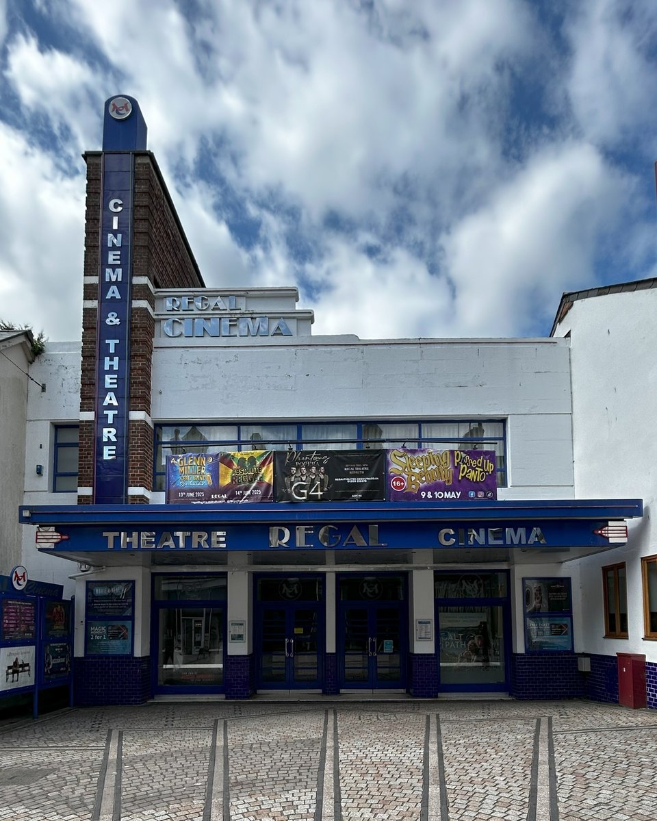 An art deco cinema front. It has a deep canopy with wide doors beneath. A ribbon of Crittall style windows is partially hidden by a banner for some shows. A fin tower spells out Cinema and theatre and a little curved sign reads Regal Cinema.