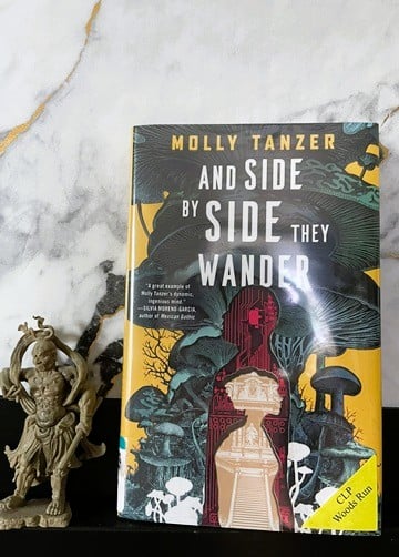 A book with a yellow background, dark green mushrooms, and two human silhouettes, looking like they were cut out of different pieces of patterned paper. It is leaning against a white marble background, and a small replica of a Japanese temple guardian statue is standing next to it