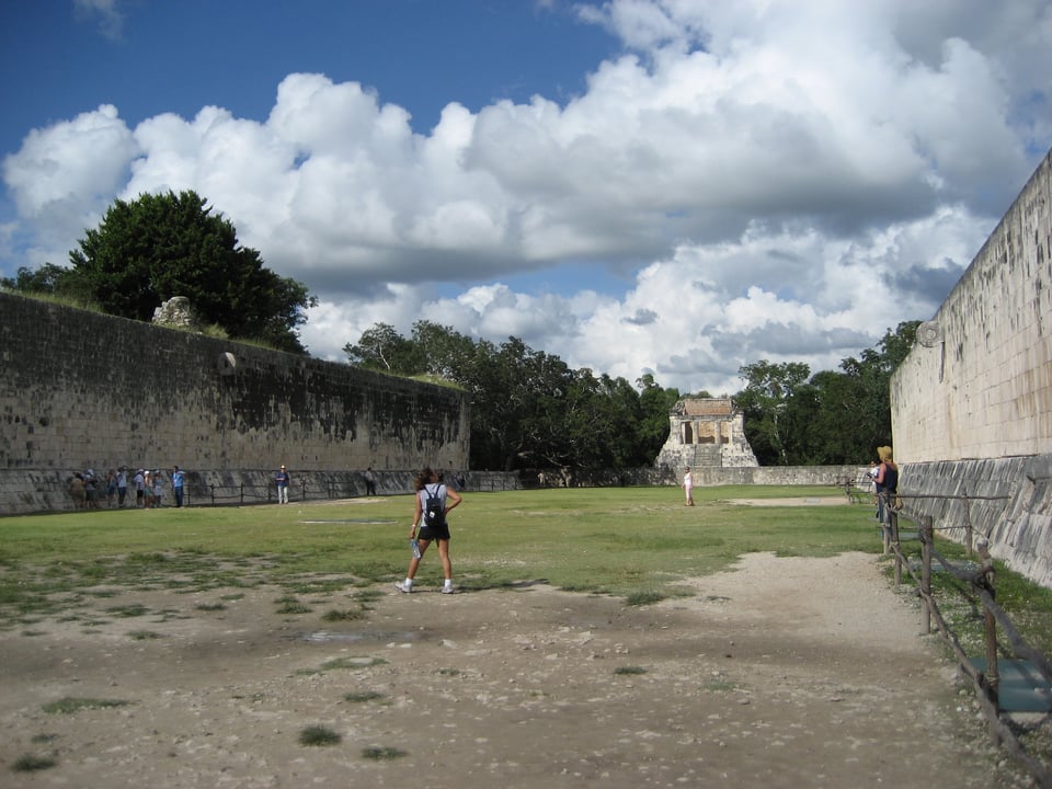 An open area with walls on either side and sparse grass. There is some kind of ruined building at the far end.