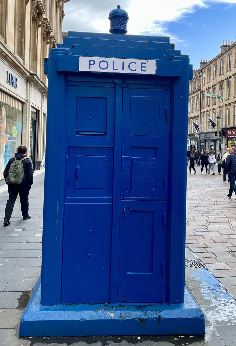 A photo of a Glasgow Police box: a blue-painted structure approximately 1.5m x 1.5m x 2.5m with panelled doors and a stepped roof with a light at its apex. A sign, centered at the bottom of the roof reads "Police".