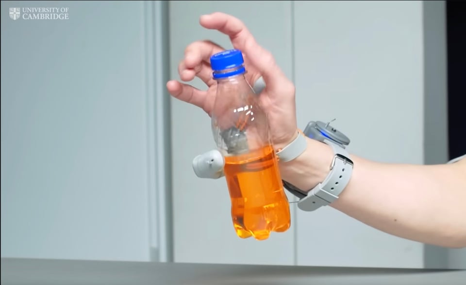 A screenshot of a woman's right arm wearing the "third thumb" strapped to her wrist. It is a grey appendage and is curled around a small plastic bottle filled with orange liquid. While the third thumb holds the bottle, her other fingers are twisting open its cap