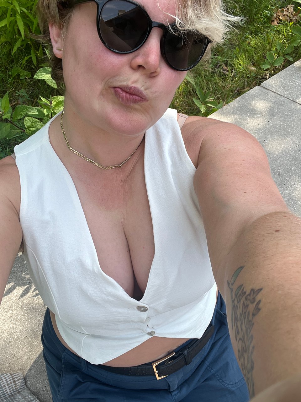 a selfie of Nola pursing her lips with bleached blonde swoopy bangs in her black sunglasses, low-cut sexy white wedding vest, gold necklace, and blue pants with black belt with gold buckle.