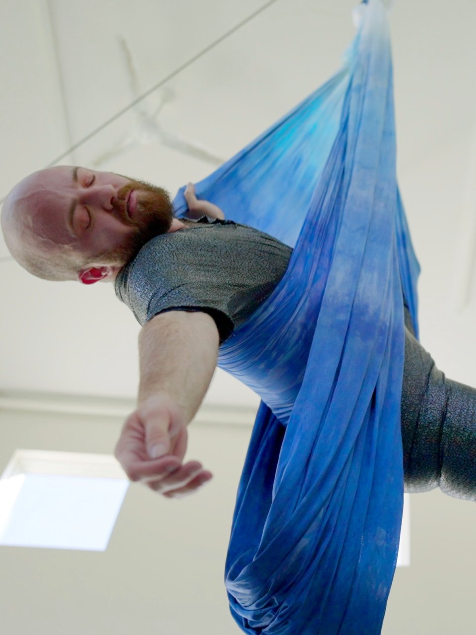 Still from the film: Looking up from below at my body held horizontal in watery-blue aerial slings, eyes closed, one arm dangling gently downward