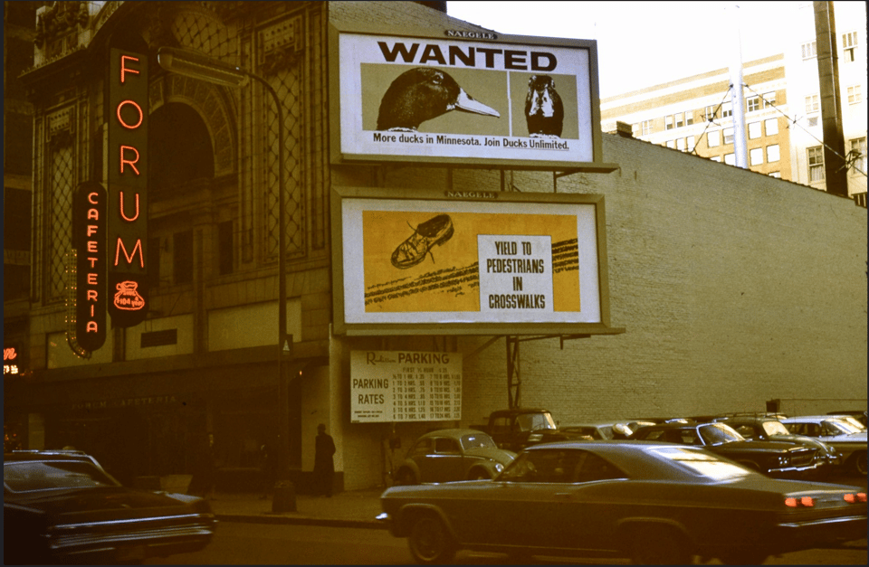 Photograph of the Forum Cafeteria, with vertical neon signs that say "FORUM" and "CAFETERIA". Two billboards are on the side of the building.