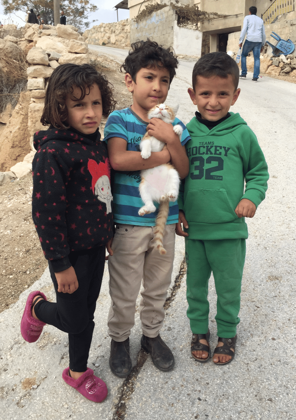 Three young children, a girl and two boys, standing on a road in front of a stone wall. The middle child is holding a cat