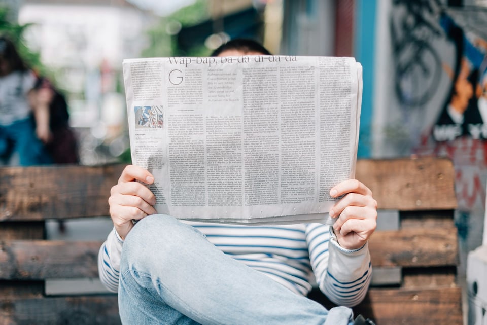 A person sitting on a bench, reading a newspaper. The newspaper covers their face and torso. Photo by Roman Kraft on Unsplash.