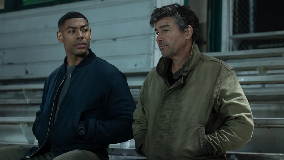 Aaron Pierre and Kyle Chandler as John Stewart and Hal Jordan in the TV show LANTERNS, seated in football bleachers in Rushville, NE, where the season has mostly been set