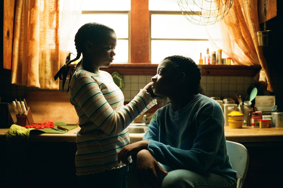 [Two girls before a window in the kitchen. One is sitting and looking up at the other one standing, who is also braiding her hair.]