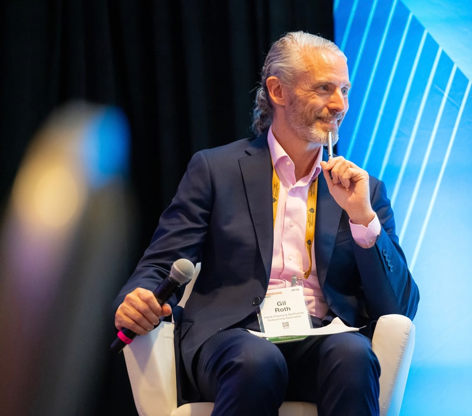 Photo of white man in a blue suit & pink shirt, sitting on a stage with a microphone in his hand, notepad on his lap, holding a pen up to his mouth as he smiles