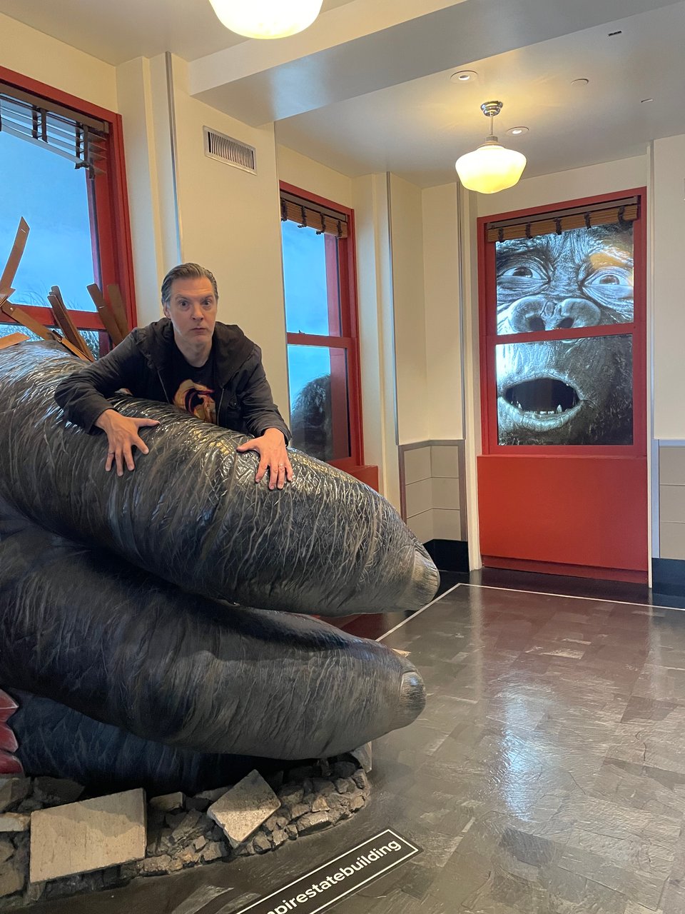 A photo op at The Empire State Building. A man appears to be held in the hand of a giant primate. The primate looks through the window, distressed at the existence of the man