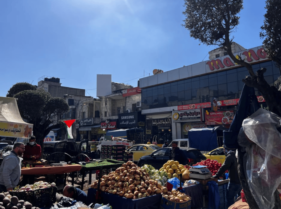 Produce for sale on a busy street, a Palestinian flag flying, and a large building on the far side of the street