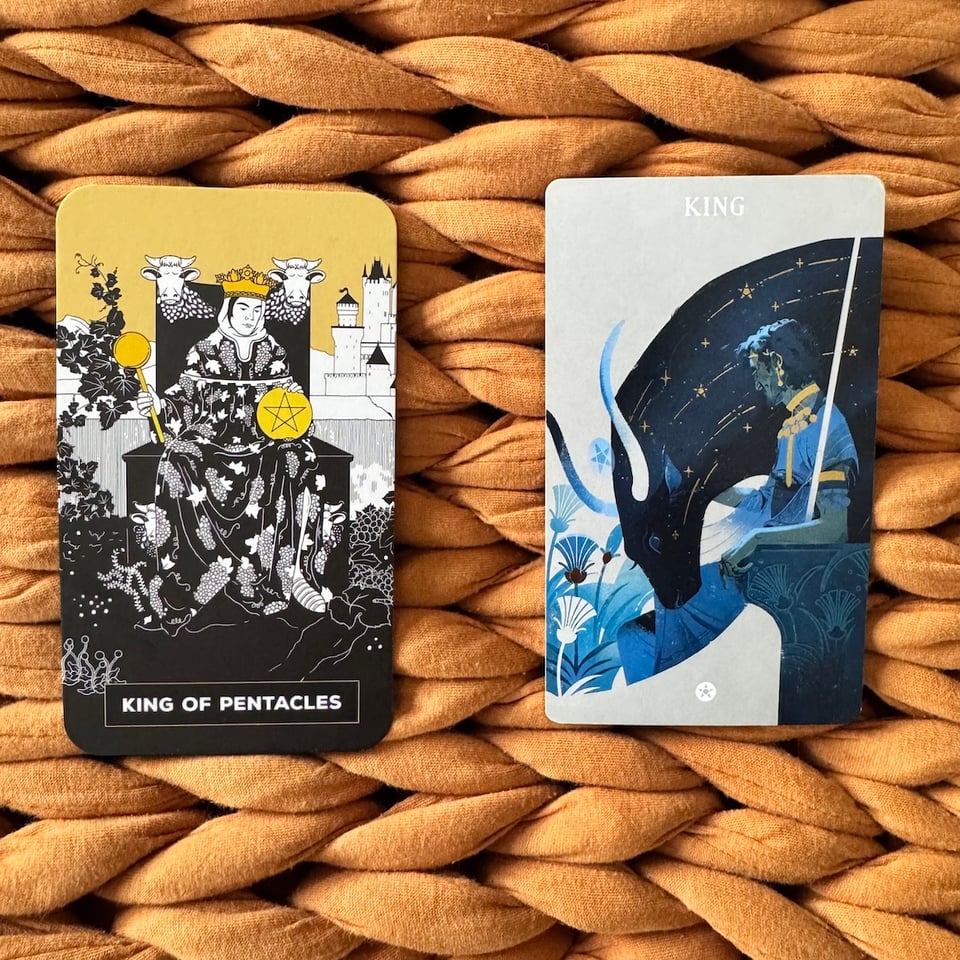Two tarot cards depicting the King of Pentacles resting on a woven beige background. The card on the left features a robed figure wearing a crown, holding a scepter and a pentacle with a bull head above the figure, the card also has a castle in the background. The card on the right showcases a dark blue figure in profile with stars, a wing and flowers in the background.