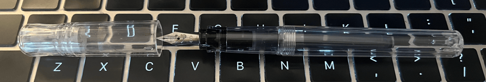 A clear plastic fountain pen sits on a laptop keyboard. The nib is metal, and it has a little face with a tongue sticking out.