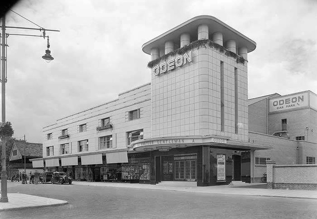 A squat ODEON cinema, faced with cream tiles. The corner tower is square, and has squat pillars supporting a flat roof. The side of the building has shops set into the ground floor, then two storeys of rooms with Crittall windows. At the ground floor, several men stand around some 1930s cars. The film showing is The Amateur Gentleman.