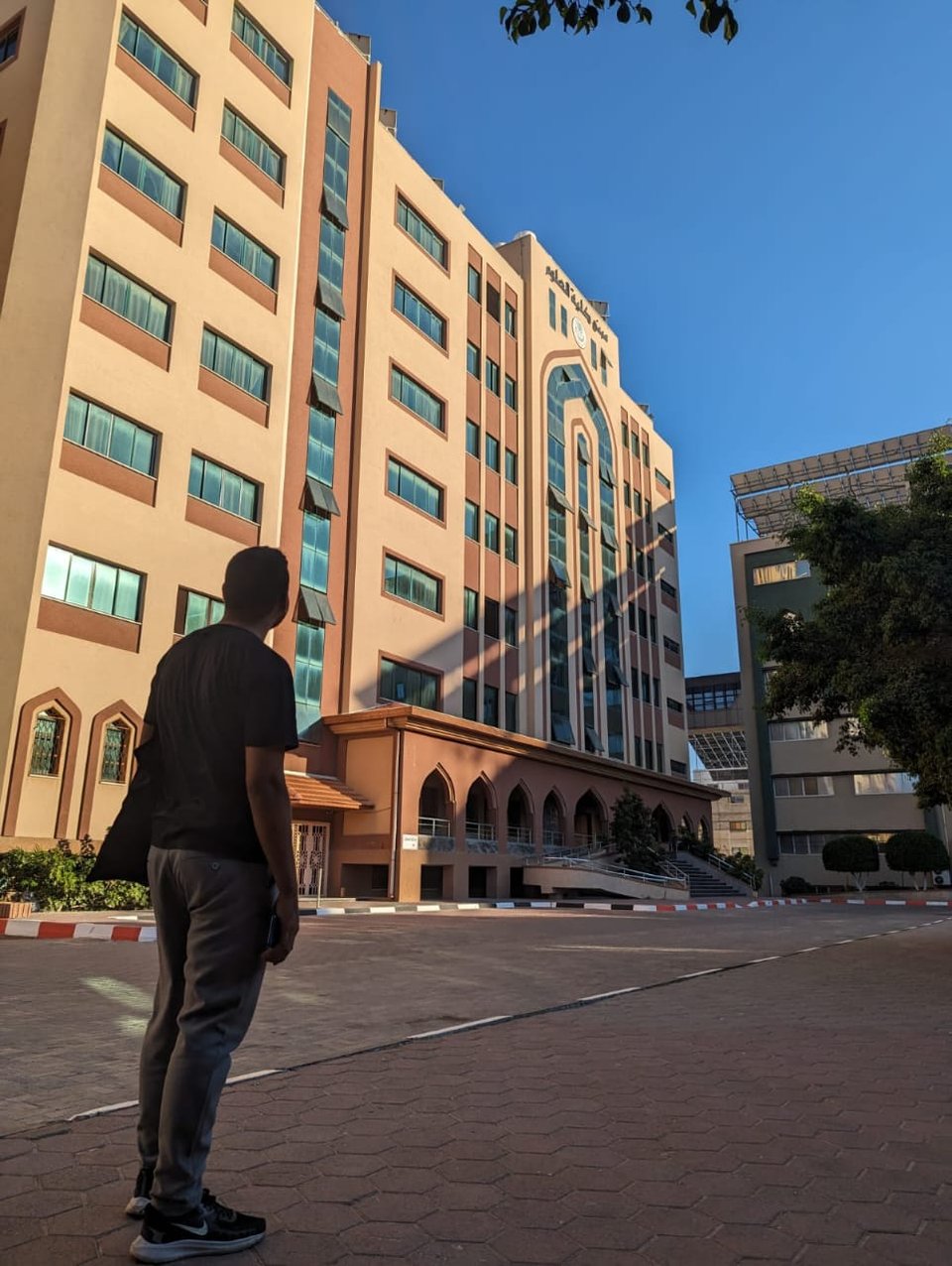 Wasim Said from behind while he stands in a roadway and looks at a tall orange-brown-beige building with some Arabic writing on it