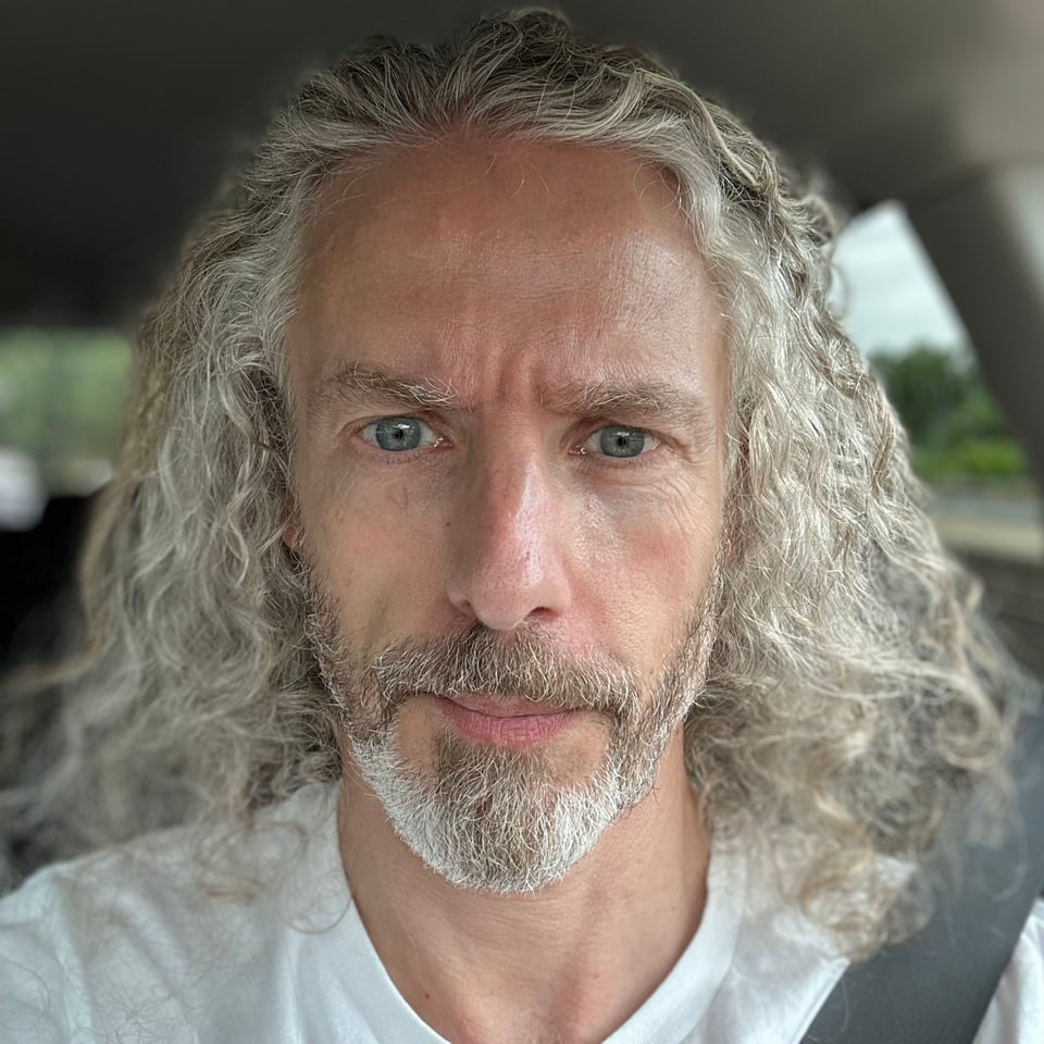 Photo of a white man with long hair and a trimmed beard, sitting in a car, seatbelt on (he was in standstill traffic; don't worry).