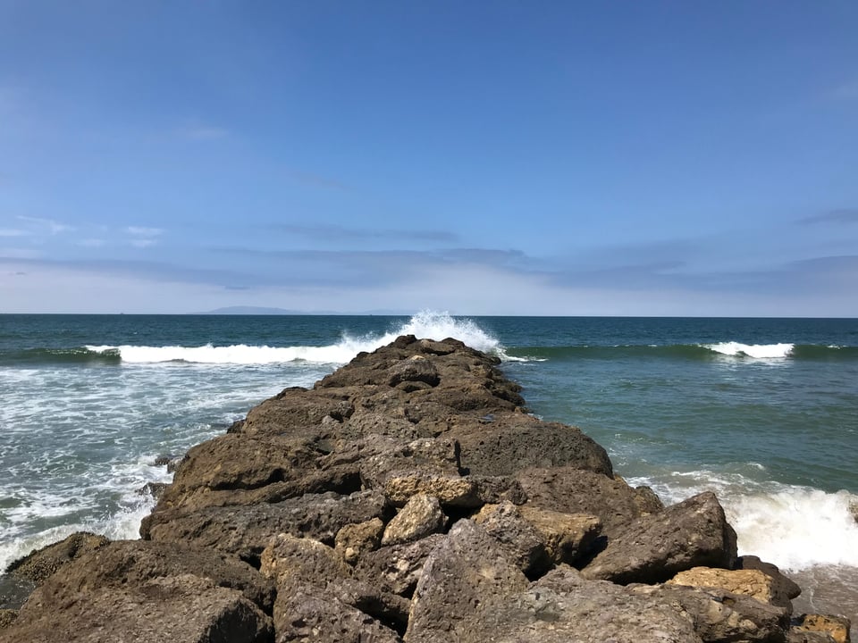A rocky breaker reaches out to the sea, a wave exploding over the edge and blue skies in the background
