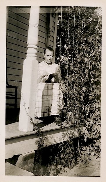 A sour faced older lady sits on a porch, doing what looks like needlepoint with either a plaid blanket or a long skirt over her legs.