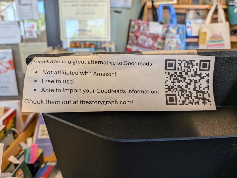 Printed sign taped to a bookstore counter promoting StoryGraph as a Goodreads alternative. The sign highlights that StoryGraph is not affiliated with Amazon, is free to use, and allows Goodreads imports, with a QR code linking to thestorygraph.com.