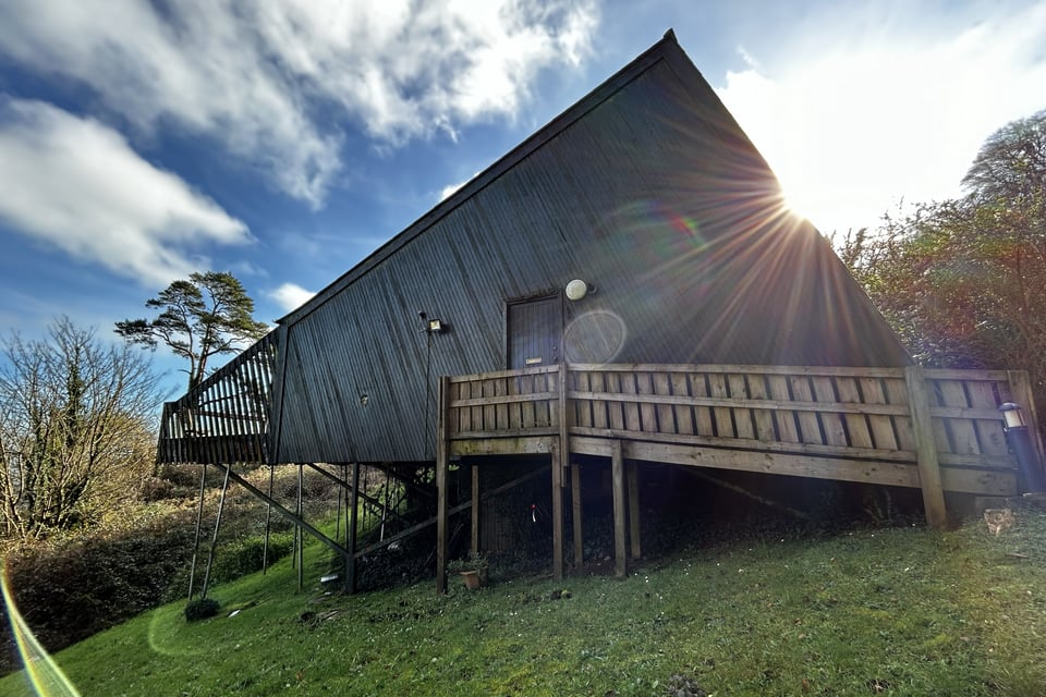 A wooden holiday lodge on a slope surrounded by trees. The lodge is wedge-shaped, with a cantilevered balcony now supported by props. The door in the side is reached by a wooden ramp.