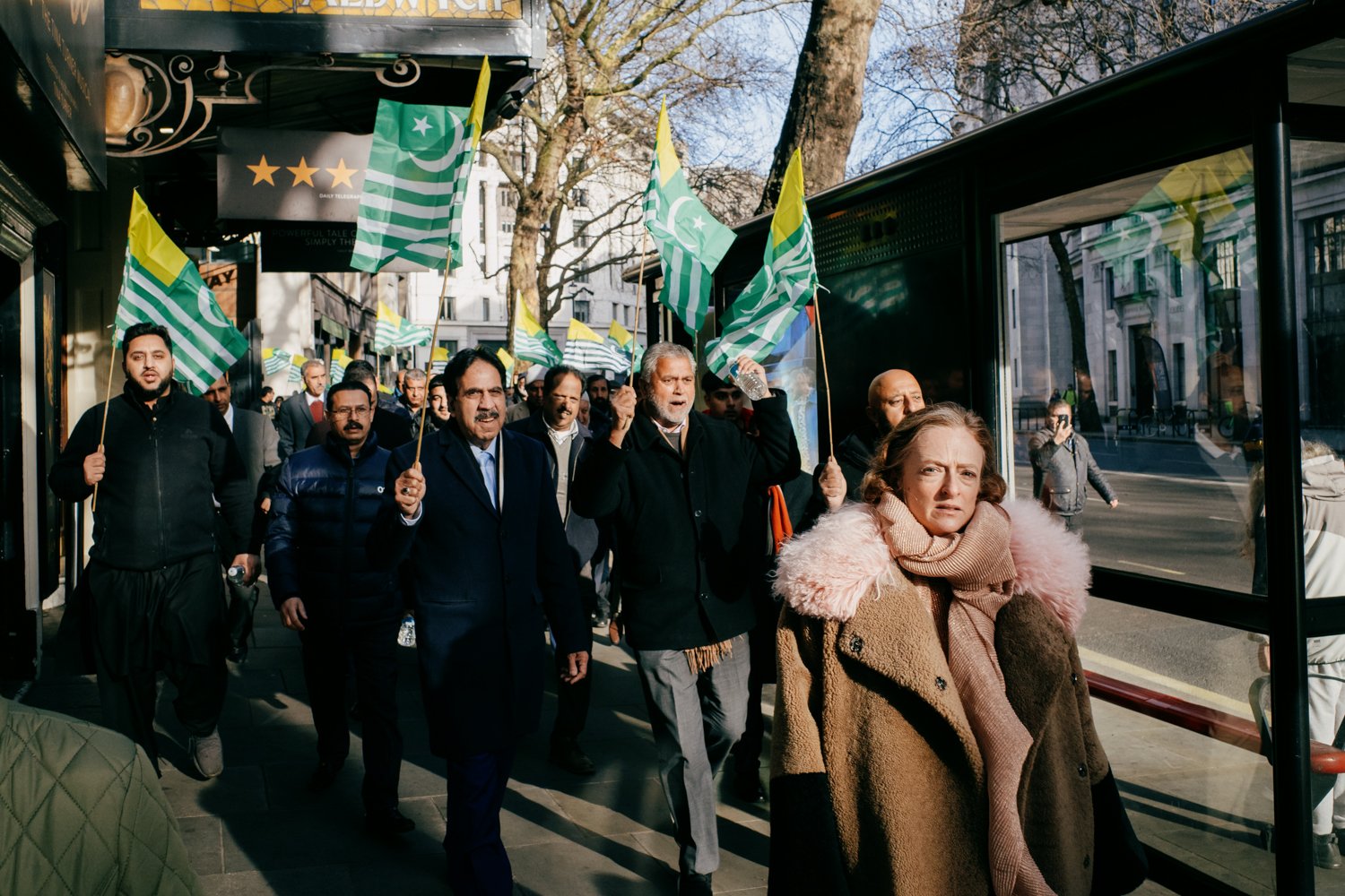 Street photo of a woman in a camel coat with pink fur trim collar looking to camera as a group of male protesters with Kashmiri flags march down the street behind her