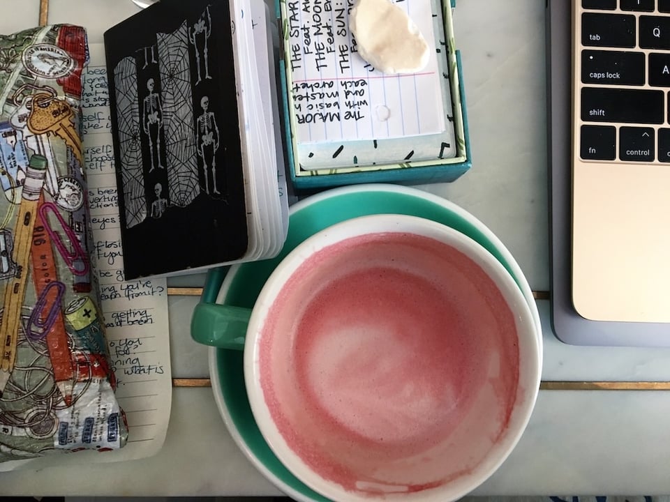 An aerial view of a pink beet latte surrounded by a partial laptop keyboard, a tarot deck with a shell on it, a tiny notebook covered in skeletons and a pen pouch covering a journal page.