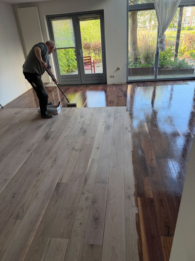 A man uses a long handled roller brush to oil newly sanded floor. The polished wood shines brightly.