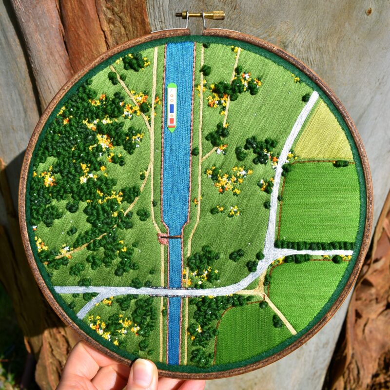 A hand holds a circular embroidery hoop showing a detailed, textured aerial landscape with fields, roads, clusters of green trees, and a bright blue canal running vertically through the center. A small embroidered boat floats on the canal, adding a pop of color against the green terrain. The stitching varies in texture and density to mimic foliage, paths, and water, giving the scene a rich, tactile depth