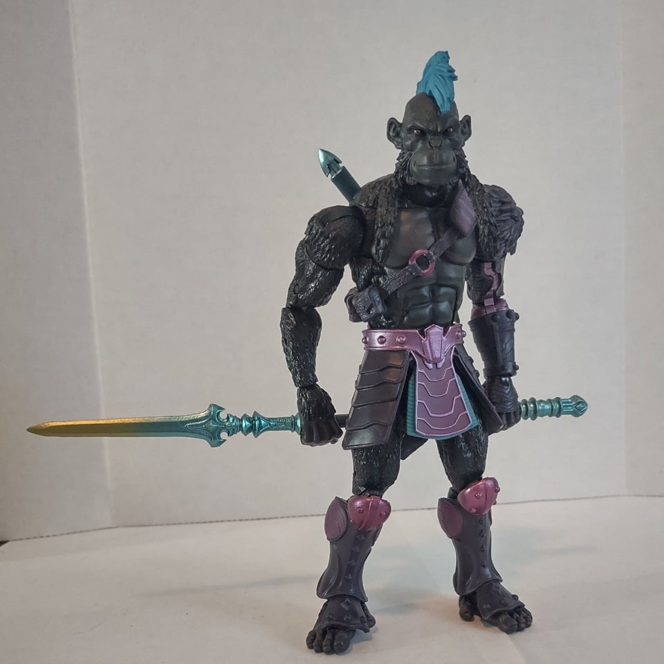 An action figure of a black furred ape-man (Blight). He has a bright blue mohawk and wears pieces of purple armor. He holds a spear in both hands behind his back