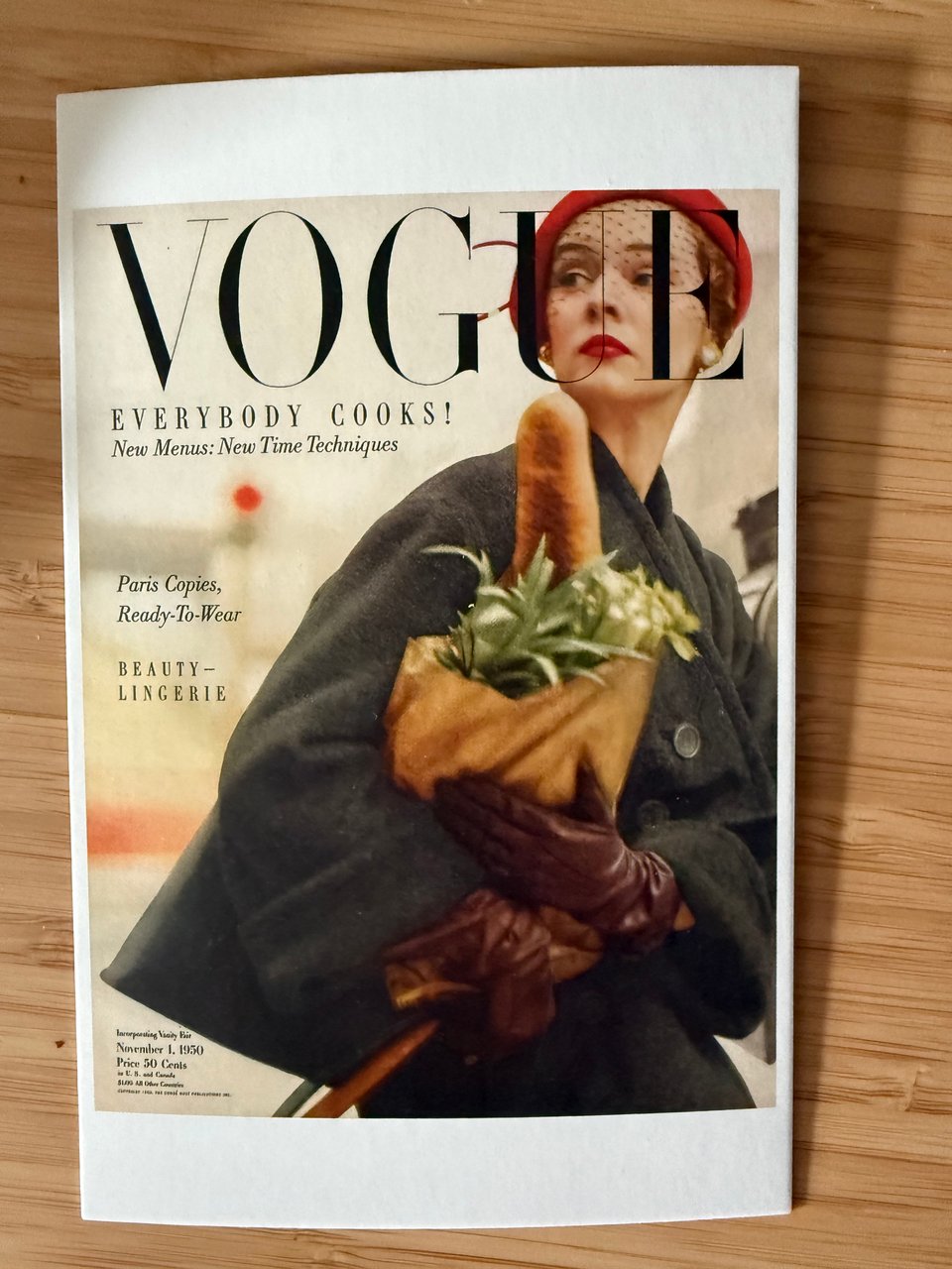 Photo of a postcard of a cover of a 1950s issue of Vogue, showing a stylish woman in an olive coat and red-brown leather gloves holding a bag of fresh produce, with a baguette sticking out toward her face and unfortunately looking very phallic