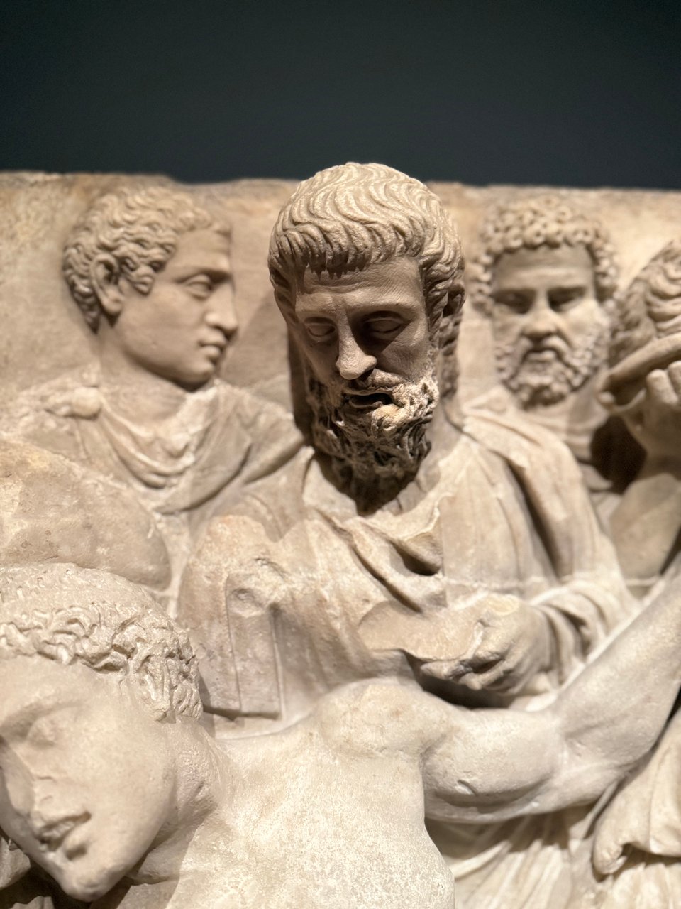 Portrait mode detail of a marble sarcophagus fragment, depicting the death of Meleager. The focus is on one of the mourners, with the overhead light casting strong shadows over his face and beard.