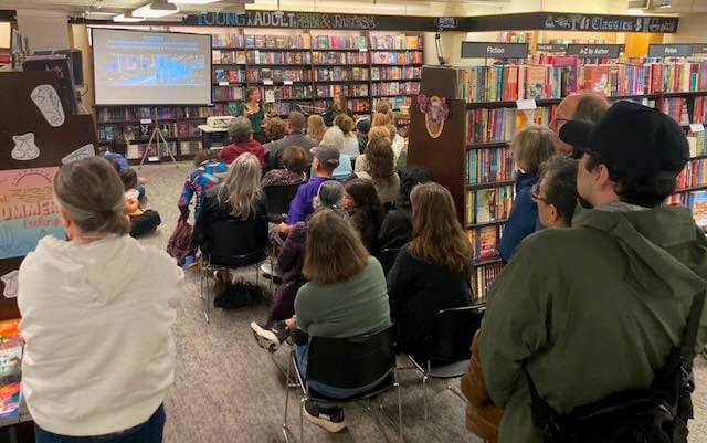 Rows of seated and standing people viewed from behind, in a bookstore lined with bookshelves; in front of them, a seated woman in green shirt in front of a projector screen.
