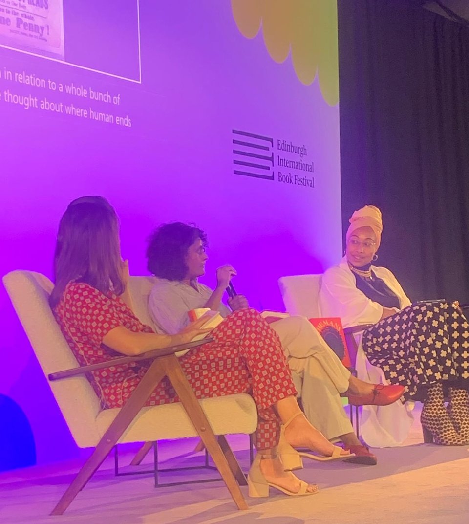 Three women on a stage. Left to right, they are wearing: a red and patterned trouser; suit and cream sandals; a pale shirt and trousers, bright red shoes, and a black face mask; black top, white robe, and black and white patterened skirt, boots, and a headwrap. In the background is a pale purple screen with “Edinburgh International Book Festival” on the right, and the corner of a printed pamphlet and part of the auto-captioning visible at the top left.