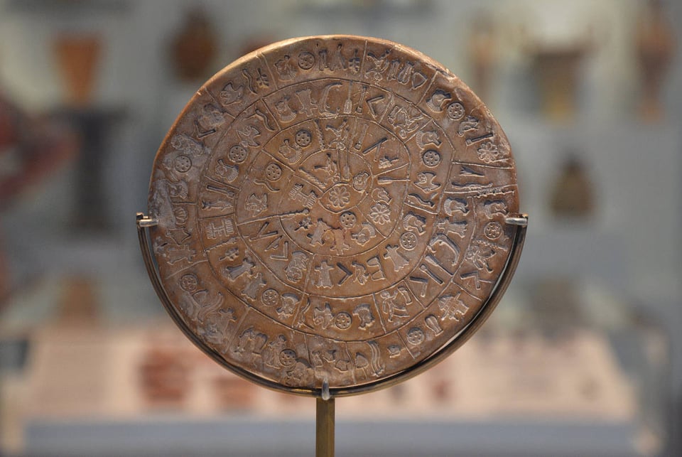 A picture of a round clay disc that is covered in indented icons, arranged in a spiral from the edge of the center. A few of the icons include small human figures; other shapes look like pieces of wood or Y shaped sticks