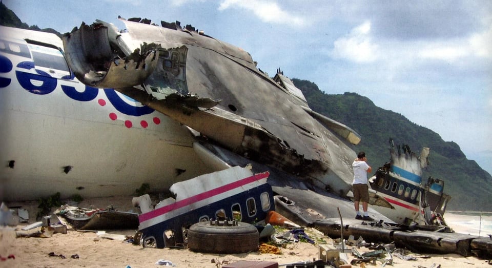 A jet plane is looking absolutely trashed on a gorgeous beach while a man stands on its wing.