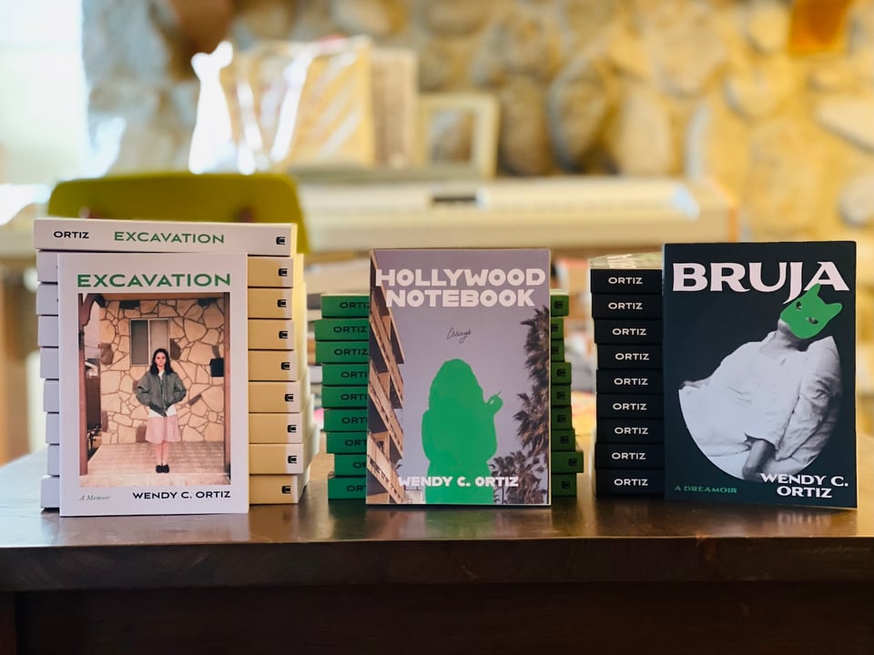 Three stacks of books, each with a copy of the book in front: Excavation, Hollywood Notebook, and Bruja, by Wendy C. Ortiz.