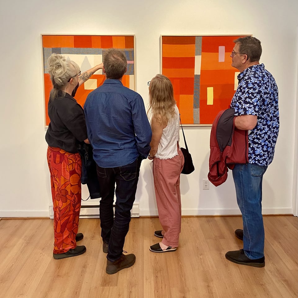 Four people stand with their backs to the camera in a gallery, looking at two large square paintings on a white wall. The works are abstract geometric compositions in deep oranges, reds, blues, and creams, with woven and striped textures. One viewer reaches up to point at the left painting. Light wood floor below.